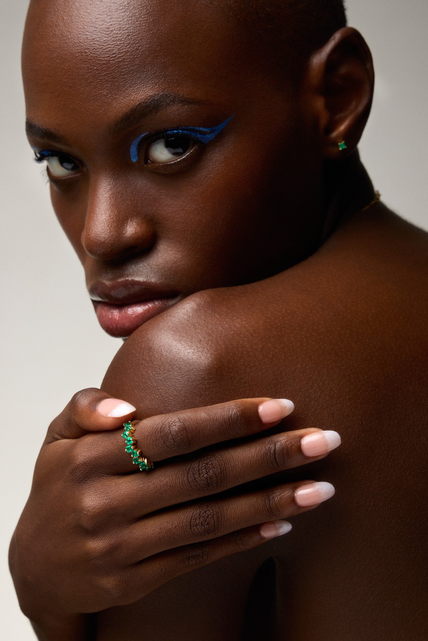 Woman with blue eyeliner and green earrings, wearing a ring, against a neutral background